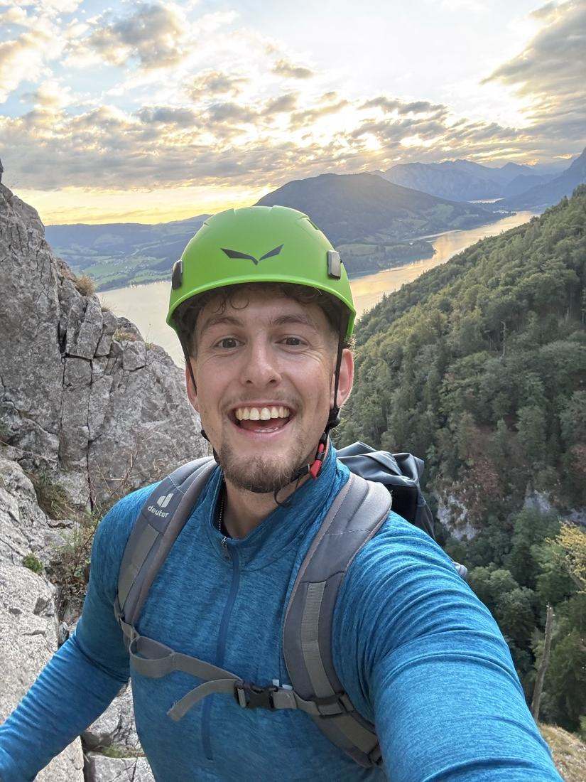 Sebi mit grünem Kletterhelm im Steig, dahinter der Mondsee im Morgenlicht.