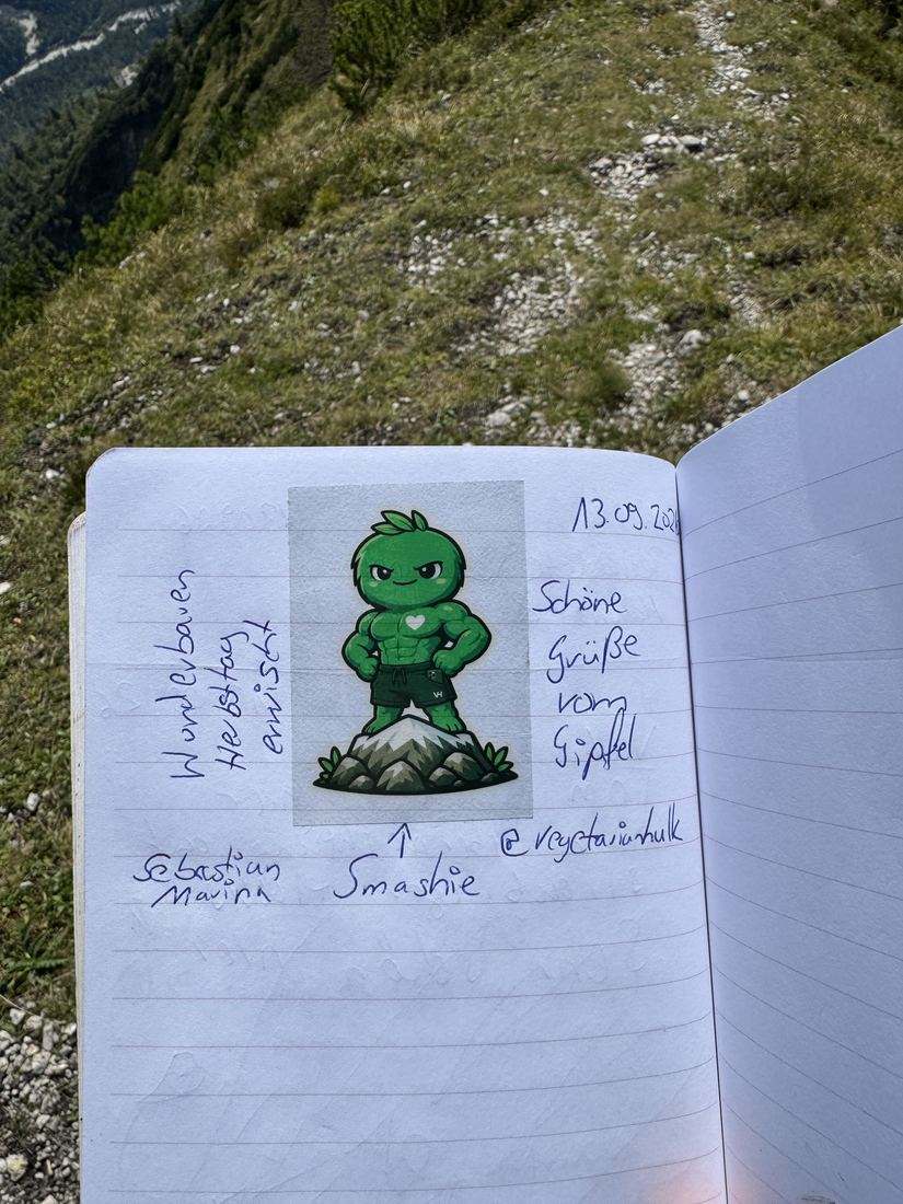 Aufgeschlagenes Notizbuch am Gipfel mit handschriftlichem Eintrag und aufgeklebtem Smashie-Sticker, daneben der Abgrund.