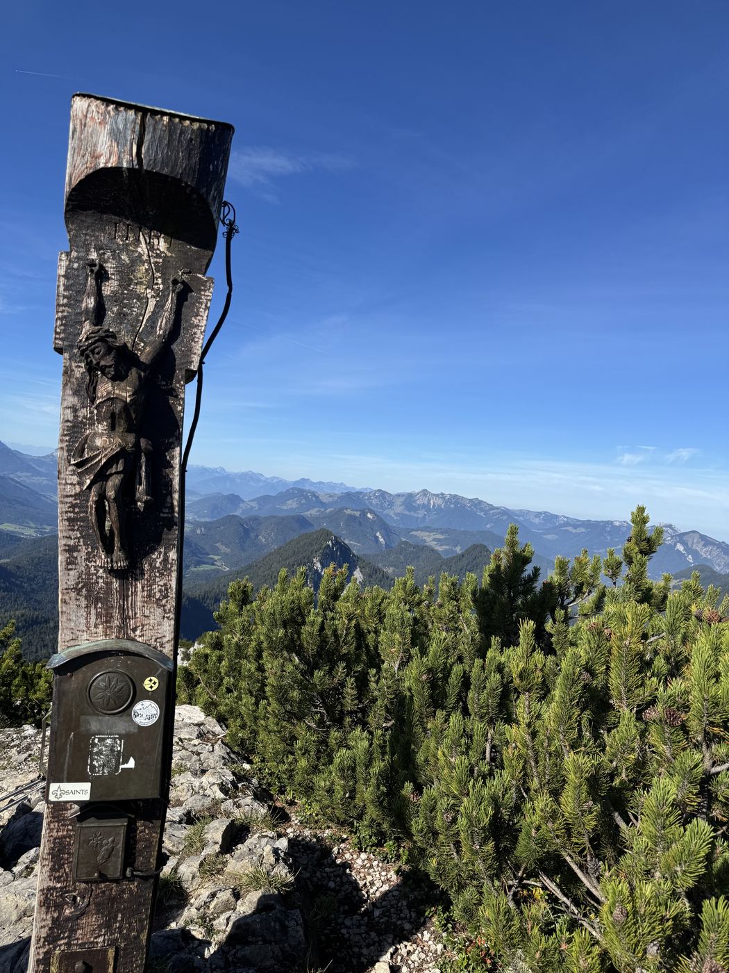 Dunkler, verwitterter Holzpfosten mit geschnitzter Figur und Gipfelkasten, dahinter die Berchtesgadener Berge.
