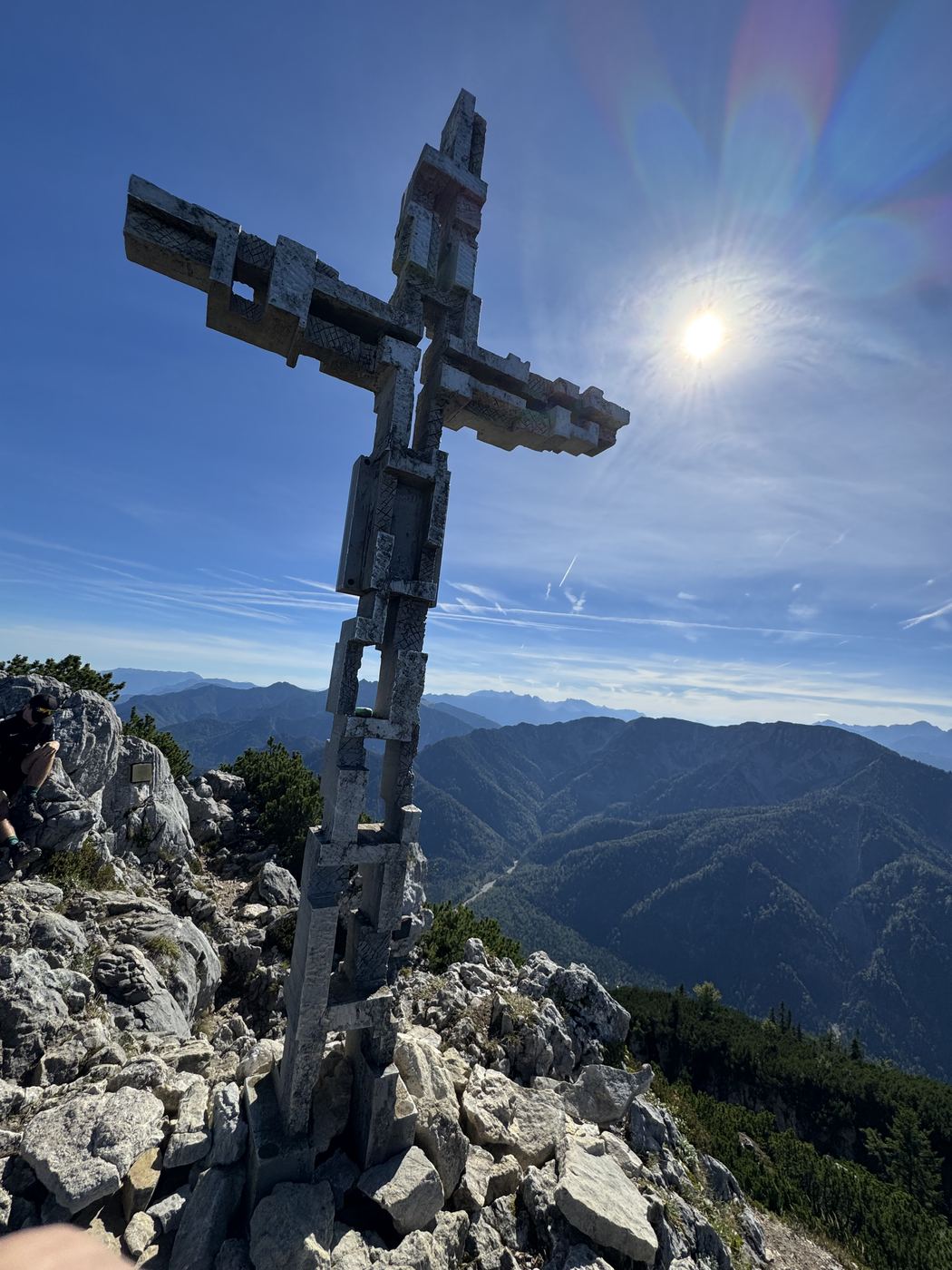 Hohes Gipfelkreuz aus verschraubtem Holz auf felsigem Grat, die Sonne steht direkt daneben.