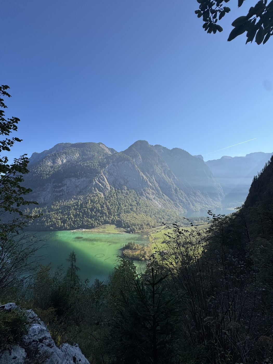 Blick vom Steig hinunter auf das türkise Seeende, darüber ein breites Felsmassiv im Gegenlicht.