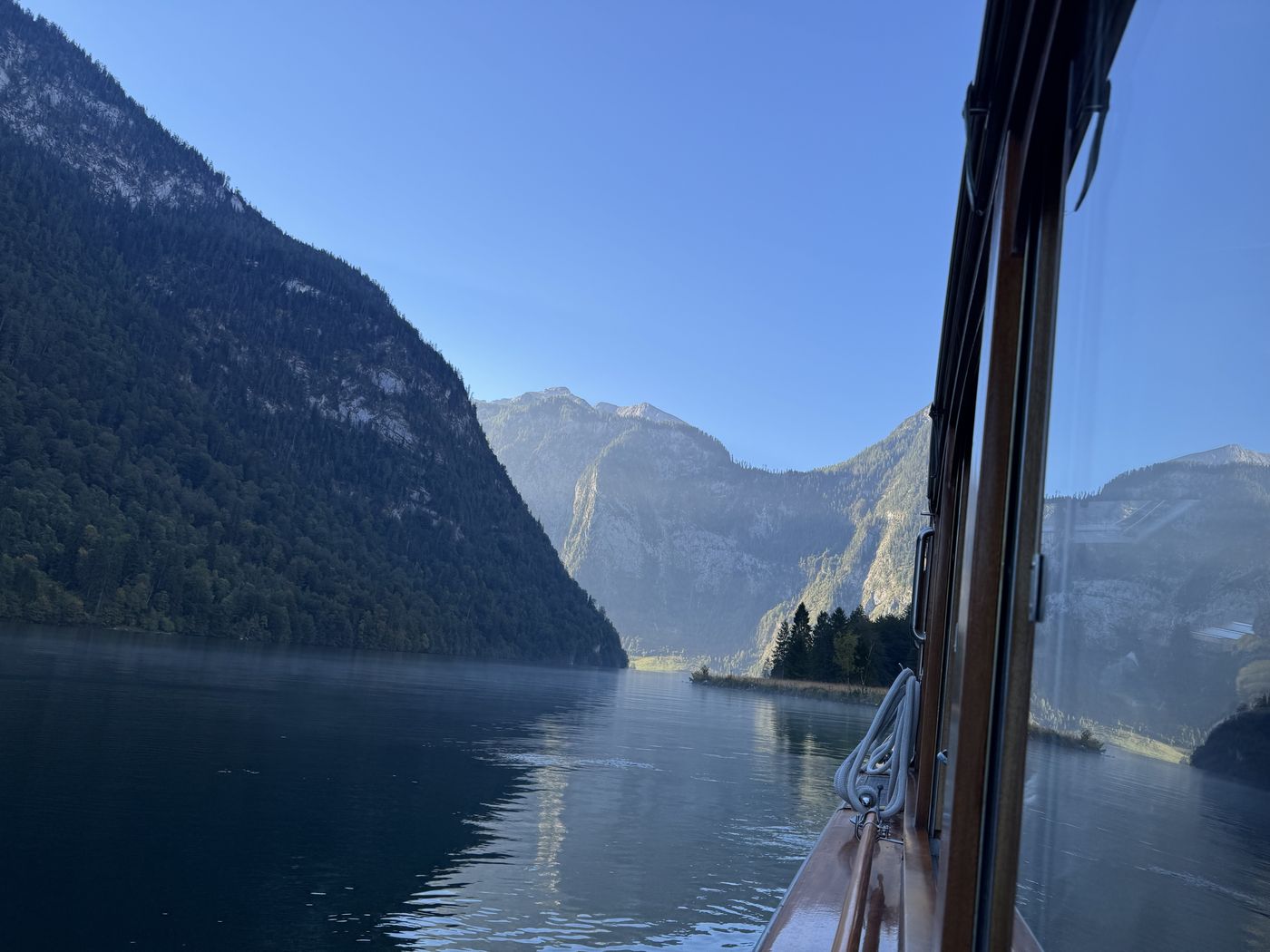 Blick vom Bug des Schiffs über den spiegelglatten Königssee, links steile Waldhänge.