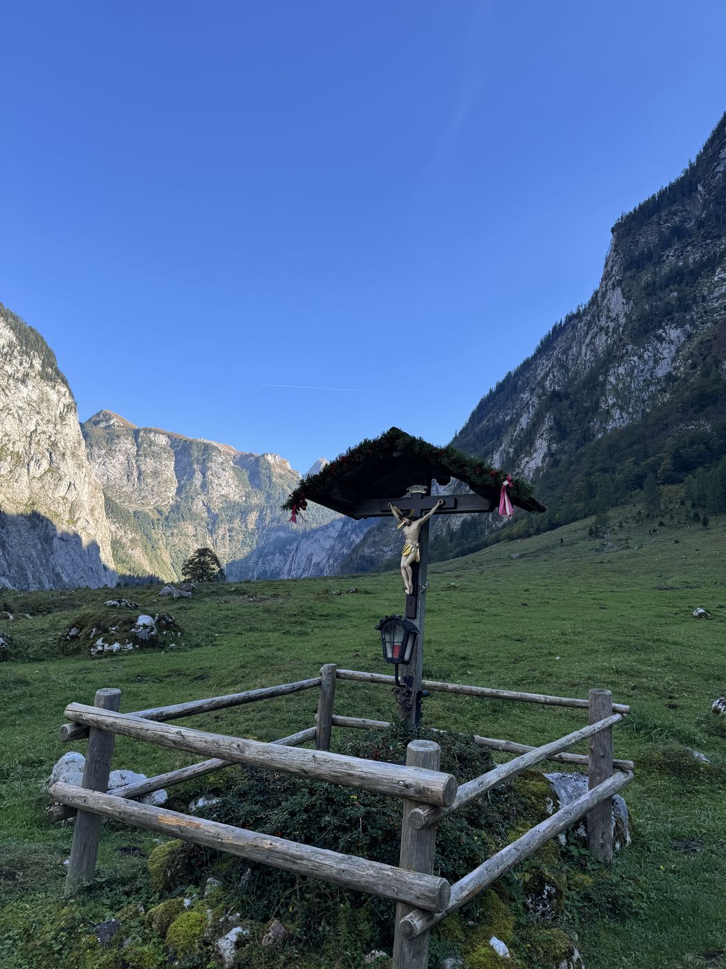 Holzkreuz mit kleinem Dach und Laterne auf einer Almwiese, eingezäunt, dahinter Felswände.