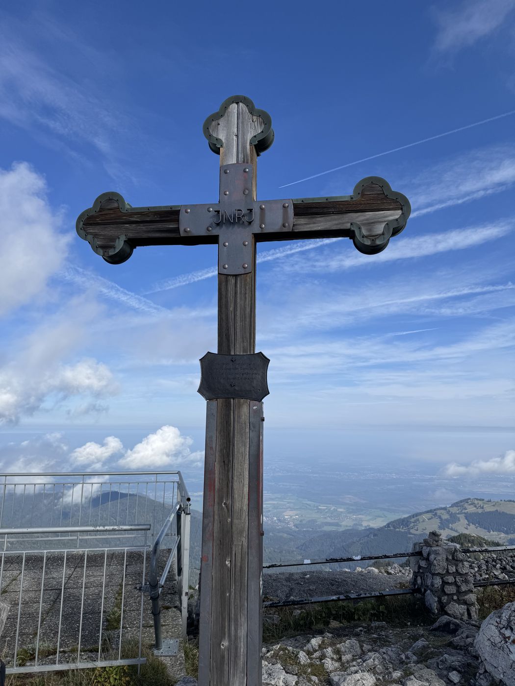 Hölzernes Gipfelkreuz mit Kleeblattenden vor blauem Himmel, unten Geländer und das Voralpenland.